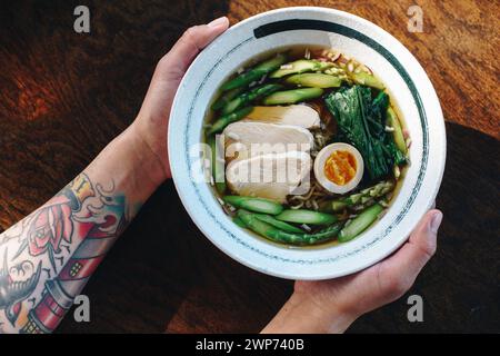 A person is holding a bowl of soup with chicken, asparagus, and egg. The soup is served in a white bowl and is placed on a wooden table Stock Photo