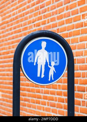 A pedestrian sign featuring a silhouette of an adult holding hands with a child is mounted against a vibrant brick wall. The bright blue circular sign Stock Photo