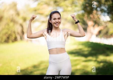 A joyful young woman in a white sports bra and leggings flexes her muscles in a sunny park Stock Photo