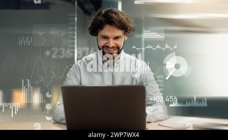 A cheerful bearded man engaging with a laptop in a modern office setting, digital graphs Stock Photo