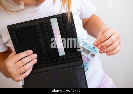 child holds black wallet in hand, kid, girl secretly takes money out of parents' wallet, pulls out euro banknotes, concept of pocket money, Personal B Stock Photo