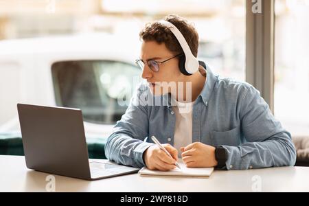 A young, curly-haired man wearing headphones writes in a notebook while working Stock Photo