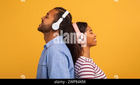 Relaxed young black couple wearing wireless headphones standing back-to-back with closed eyes Stock Photo
