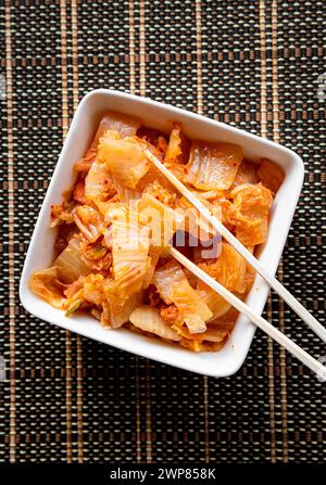 Selective focus on bamboo chopsticks with fermented kimchi cabbage salad and white food bowl on brown background. Stock Photo