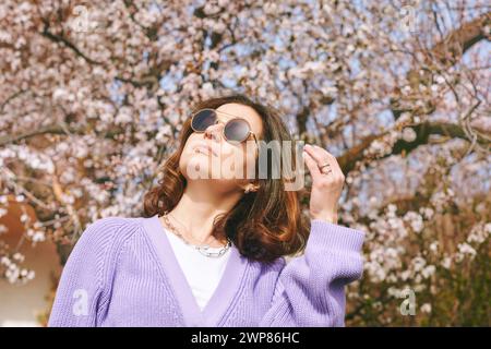 Outdoor fashion portrait of beautiful woman posing next to blooming spring tree, wearing purple knitted cardigan Stock Photo