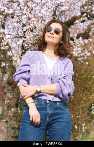 Outdoor fashion portrait of beautiful woman posing next to blooming spring tree, wearing purple knitted cardigan Stock Photo