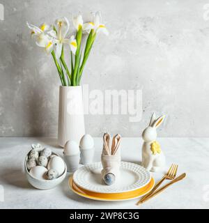 White monochrome Easter table. Natural colors eggs, figured plates, ceramic bunny, napkin as bunny muzzle, bouquet of white Iris flowers on table. Cop Stock Photo