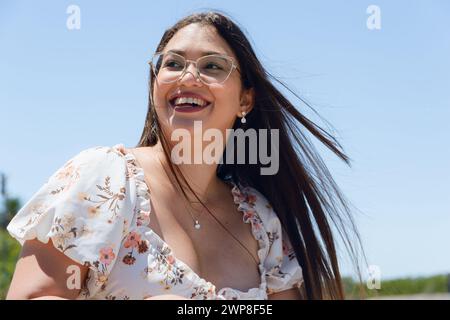 young Latin woman of Venezuelan ethnicity and white skin, with glasses, white flowered dress and long hair blown by wind is smiling happily outdoors l Stock Photo