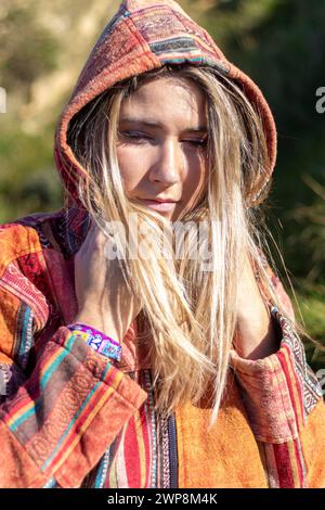 woman with long blonde hair is wearing a colorful jacket with a hood. She is looking at the camera with a thoughtful expression Stock Photo