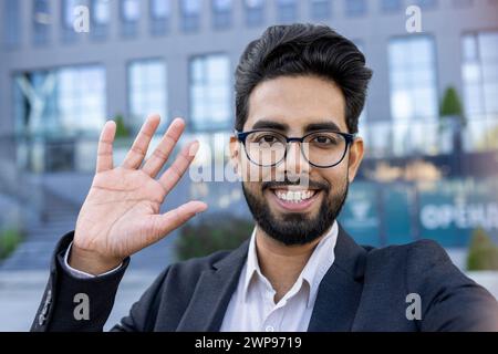 Portrait of a cheerful Indian businessman waving his hand in greeting. Standing outdoors with a bright smile, he portrays a casual yet professional demeanor. Represents multicultural business environments. Stock Photo