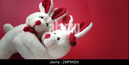Two white plush rabbits cuddling. Dark red background. Children's toys are white. Greeting card on Valentine's Day. Mutual love and affection Stock Photo