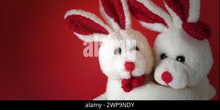 Two white plush rabbits cuddling. Dark red background. Children's toys are white. Greeting card on Valentine's Day. Mutual love and affection Stock Photo