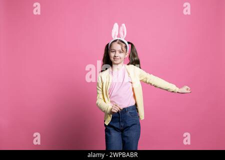 Small carefree girl acting positive with bunny ears on camera, lovely child fooling around and dancing against pink background. Little child getting excited and joyful about easter holiday. Stock Photo