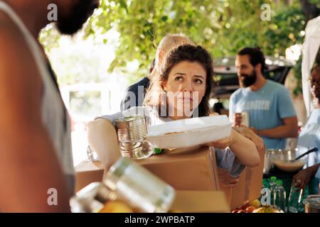 Portrait shot of poor and needy caucasian woman carrying her donation box and canned goods from food bank. Image showing the underprivileged and homeless people receiving support from charity group. Stock Photo