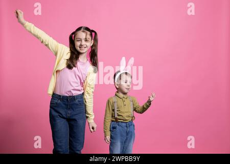Happy young siblings dancing around being funny in studio, feeling joyful and excited about easter festivity and presents. Cute brother and sister showing fun dance moves against pink background. Stock Photo