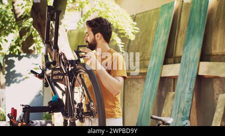 Caucasian male cyclist mending his own bicycle in the yard doing annual outdoor vehicle maintenance using professional equipment. Examining and repairing bike gear as summer hobby. Stock Photo