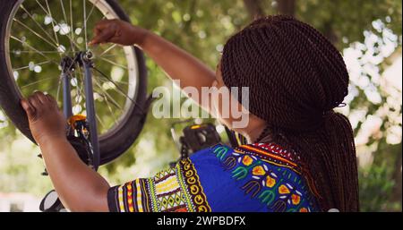 Healthy black woman mending her own bicycle using specialized equipment from toolkit in yard. Dolly zoom-in shot. Female african american cyclist repairing front bike wheel outdoors. Stock Photo