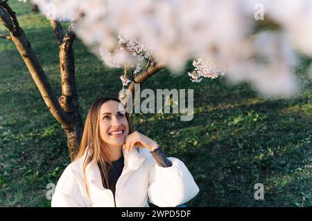 Beautiful woman in a white jacket sits under blooming tree in city park in spring Stock Photo