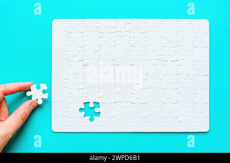 Top view of a hand placing the final piece of a blank rectangular jigsaw puzzle set on a pristine blue background. Stock Photo