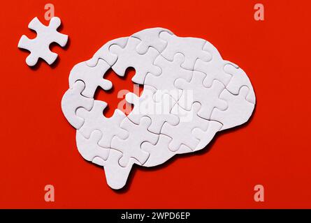 Human brain-shaped puzzle missing a crucial piece lying beside it on a red backdrop. Mental health issues related concept. Stock Photo