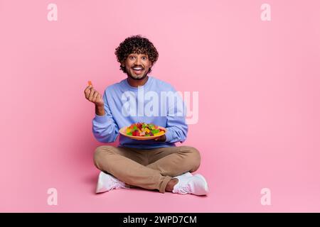 Photo of cheerful funny guy sitting enjoying delicious candies snack relax break pause isolated on pink color background Stock Photo