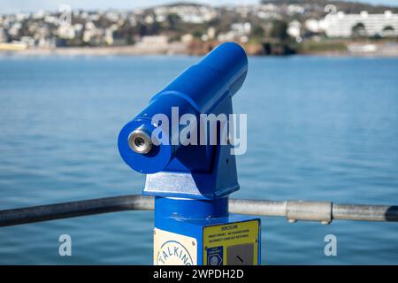 A blue viewing telescope looks out over a calm blue sea towards land at a popular holiday destination in Devon Stock Photo