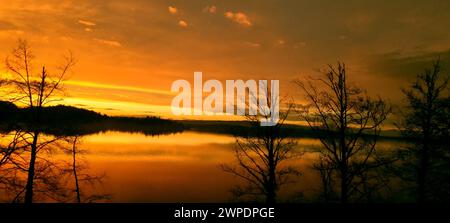 The sun sets behind trees, casting warm light on a tranquil lake Stock Photo
