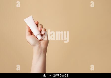 Female hand holding light tube with Foundation or cosmetic cream on beige background. Cosmetic beauty product branding mockup. Copy space Stock Photo