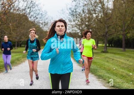 An active group of women in various colors of sportswear, from blue to neon green, run together along a park's gravel pathway, enjoying the camaraderi Stock Photo