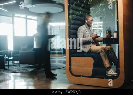 Businessman working in a busy privacy booth, having a video call on his laptop. The young entrepreneur is fully engaged in the virtual meeting, discus Stock Photo