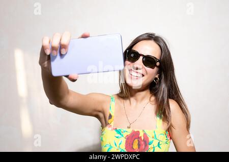 A cheerful young woman with long hair and sunglasses captures a joyful moment with her smartphone, taking a selfie while wearing a colorful summer dre Stock Photo