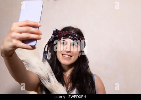 A vivacious young woman adorned with a stylish headscarf smiles as she captures a selfie using her smartphone, exuding a sense of joy and contemporary Stock Photo