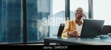 Businesswoman in a modern office, using her laptop and speaking on the phone. She is confident and successful, dressed in corporate attire. Woman comm Stock Photo