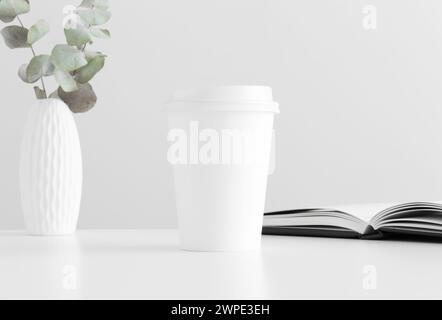 Coffee paper cup mockup with eucalyptus in a vase and a notebook on a white table. Stock Photo