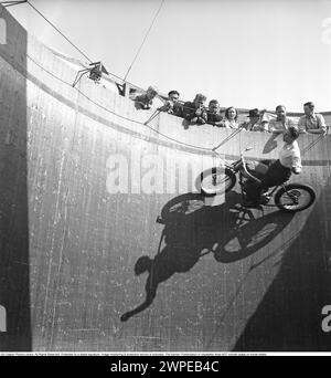 Wall of death 1949. The wall of death, motordrome, velodrome or well of death is a carnival sideshow featuring a silo- or barrel-shaped wooden cylinder, typically ranging from 20 to 36 feet (6.1 to 11.0 m) in diameter and made of wooden planks, inside which motorcyclists travel along the vertical wall and perform stunts, held in place by friction and centrifugal force. The original wall of death was in 1911 on Coney Island in the United States. Pictured a motorcyclist in high speed in the velodrome, showing off, with his hands in the air, not on the handlebars as he should. Sweden 1949. Conard Stock Photo