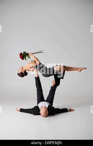 flexible woman in black attire holding red roses and balancing on feet of dancing partner on grey Stock Photo