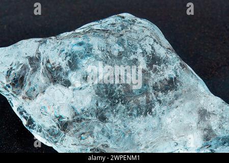 Close-up of a clear, textured ice chunk that looks like a diamond on a dark surface, highlighting natural patterns and bubbles within the ice. Locatio Stock Photo