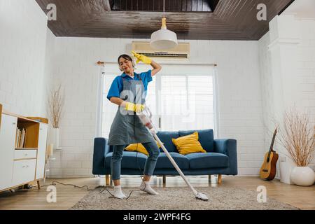 Happy maid's cleanup melody, Asian woman uses mop as mic singing dancing. Fun-filled service with Stock Photo