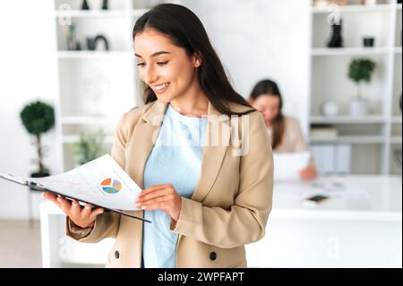 Clever pretty indian or arabian business, company executive, financial manager, stand in a modern office, holding and studying documents, analyzing financial results, smiling Stock Photo