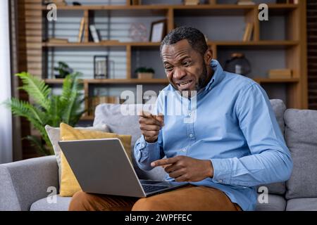A middle-aged man sits on his couch, looking perplexed and frustrated while trying to figure out his laptop. Stock Photo