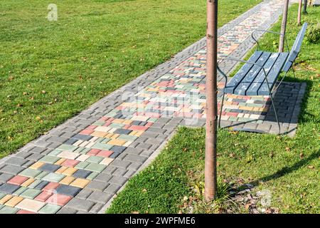 Colorful paved road across green lawn with empty bench on sunny day. Pathway and seat in summer park. Tiled pathway across public garden Stock Photo