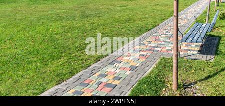 Colorful paved road across green lawn with empty bench on sunny day. Pathway and seat in summer park. Tiled pathway across public garden Stock Photo