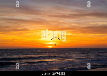 Beautiful and peaceful solo parasailer paramotoring over the ocean at orange and yellow sunset with surfers in the Pacific Ocean, Stock Photo