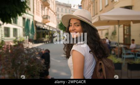 Walking outdoor cheerful smiling stylish Indian Arabian woman lady female tourist traveler follows back spinning turning have fun looking camera Stock Photo