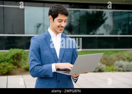 A focused Hispanic businessman in a sharp blue suit reviews data on his laptop while standing outdoors, representing the modern entrepreneur ability to work from anywhere. Stock Photo
