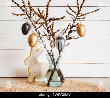 Tree branches in vase with black and bronze colored Easter eggs hanging on string, white rustic wood board background. Minimal home Easter decoration. Stock Photo