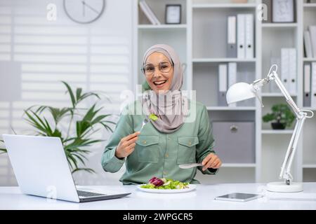 A joyful Muslim woman wearing a hijab eats a fresh salad at her desk, taking a break from work with her laptop and lamp beside. Stock Photo