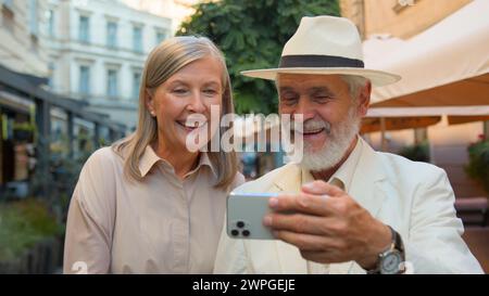 Happy smiling Caucasian senior elderly couple walking taking selfie laughing talking gadget mobile phone together city street outdoors. Old man woman Stock Photo