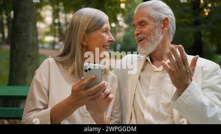 Happy elderly Caucasian couple retired 70s family married aged spouses having fun using mobile phone online call app outdoors city nature park Stock Photo