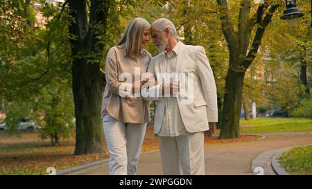 Happy healthy elderly senior mature Caucasian grandparents married man woman family couple walking holding hands outside city park telling stories Stock Photo